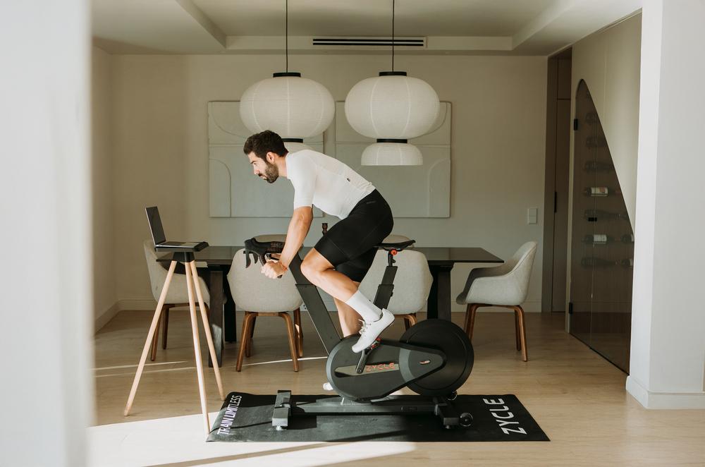 Una persona pedalea en una ZBike Infinity sobre una esterilla junto a la mesa del comedor y un soporte para el ordenador.